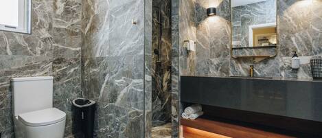 Un baño con pared de mármol, un inodoro, un cubo de basura, un lavamanos con espejo y un perchero para toallas.