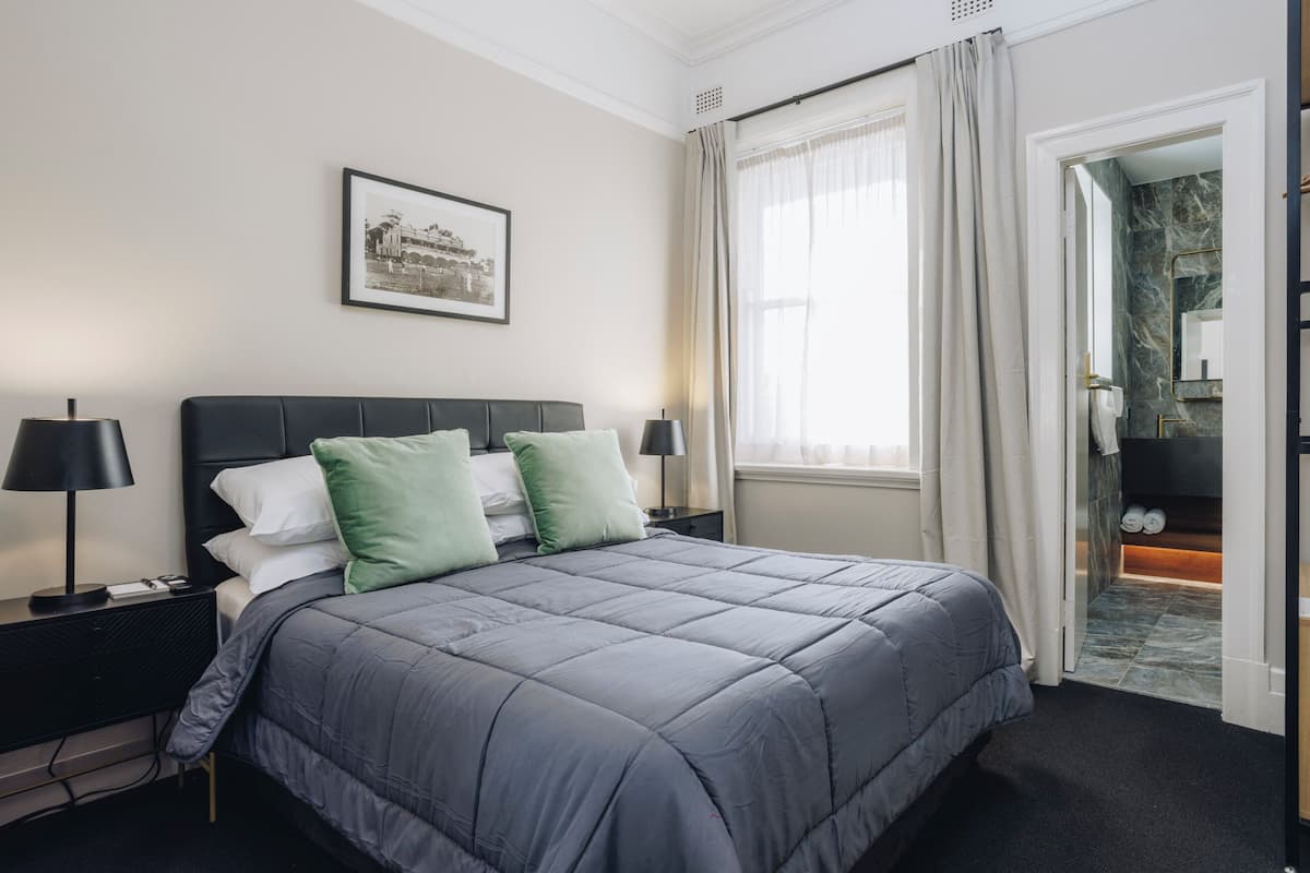 A neatly arranged hotel room with a large bed, a bedside table with a lamp, a framed picture on the wall, and a bathroom visible through an open door.