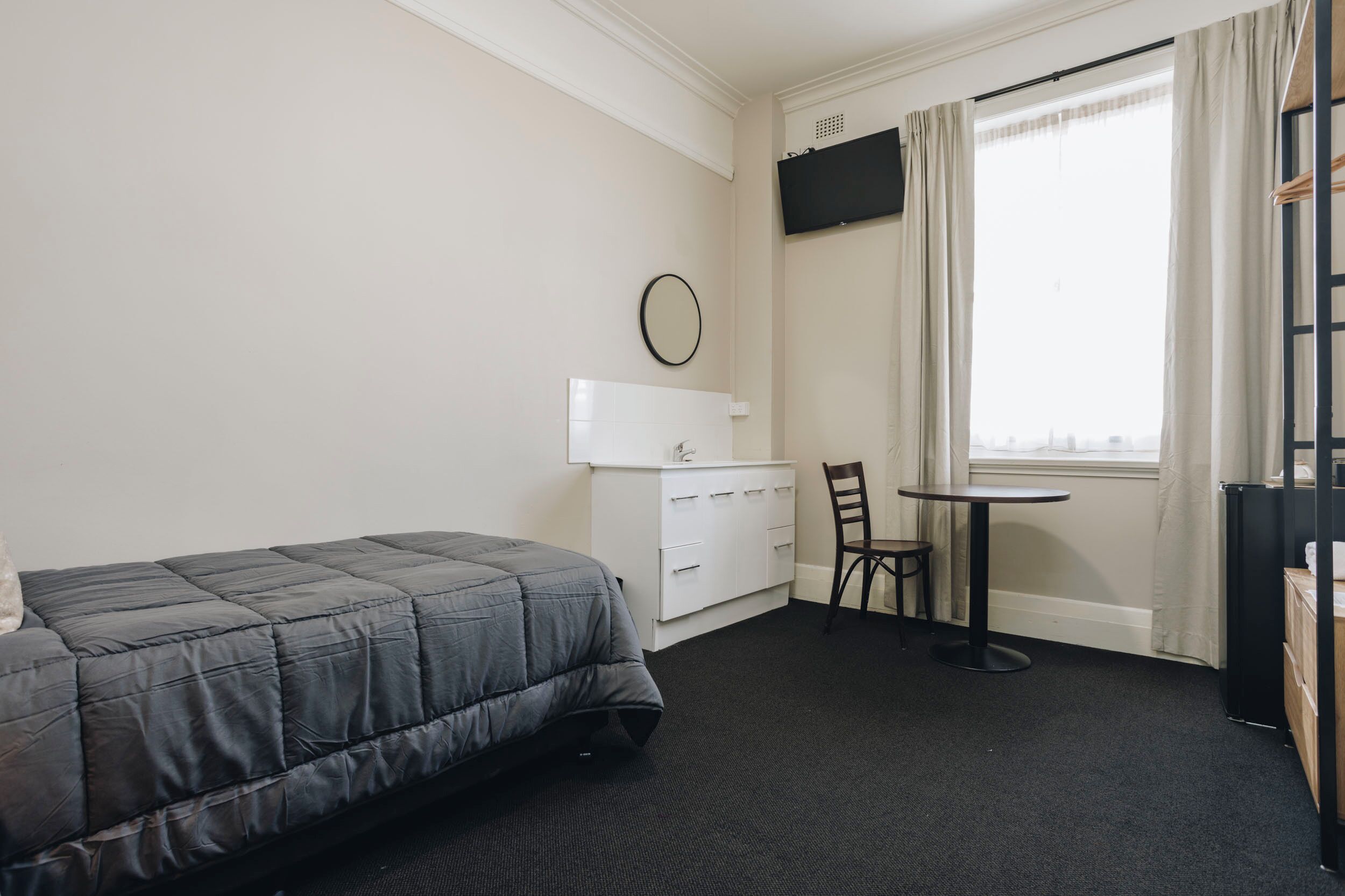 A hotel room with a bed, a small desk with a chair, a television mounted on the wall, and a window with curtains.