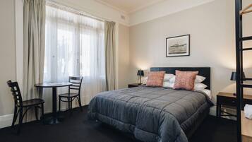 A hotel room with a large bed, a desk, two chairs, a window with curtains, a nightstand with a lamp, and a framed picture on the wall.