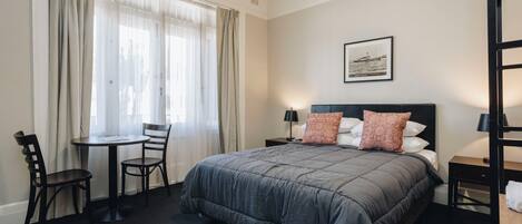 A hotel room with a large bed, a desk, two chairs, a window with curtains, a nightstand with a lamp, and a framed picture on the wall.