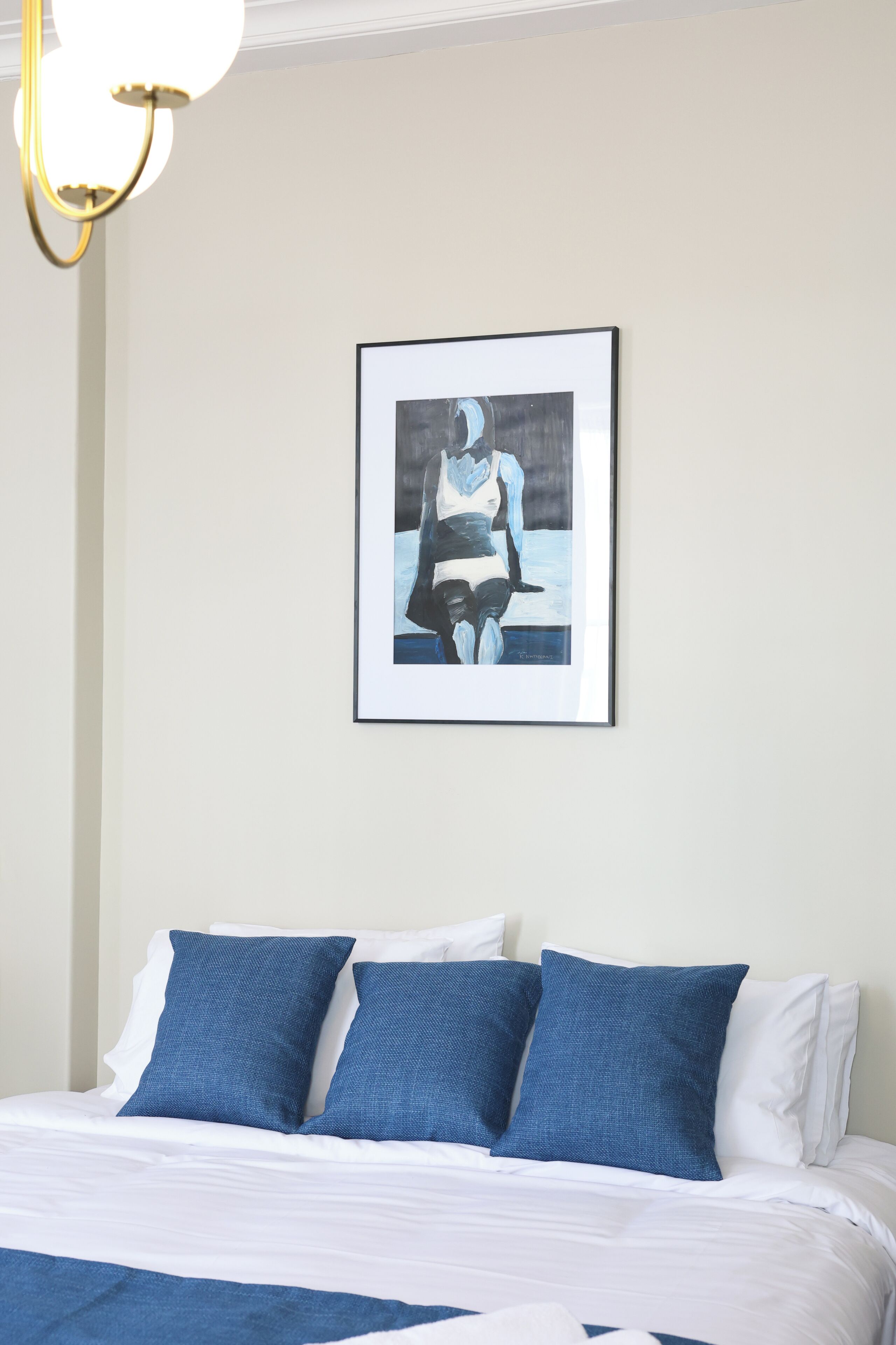 Una cama bien tendida con ropa de cama azul y blanca, un cuadro en la pared y una lámpara sencilla.