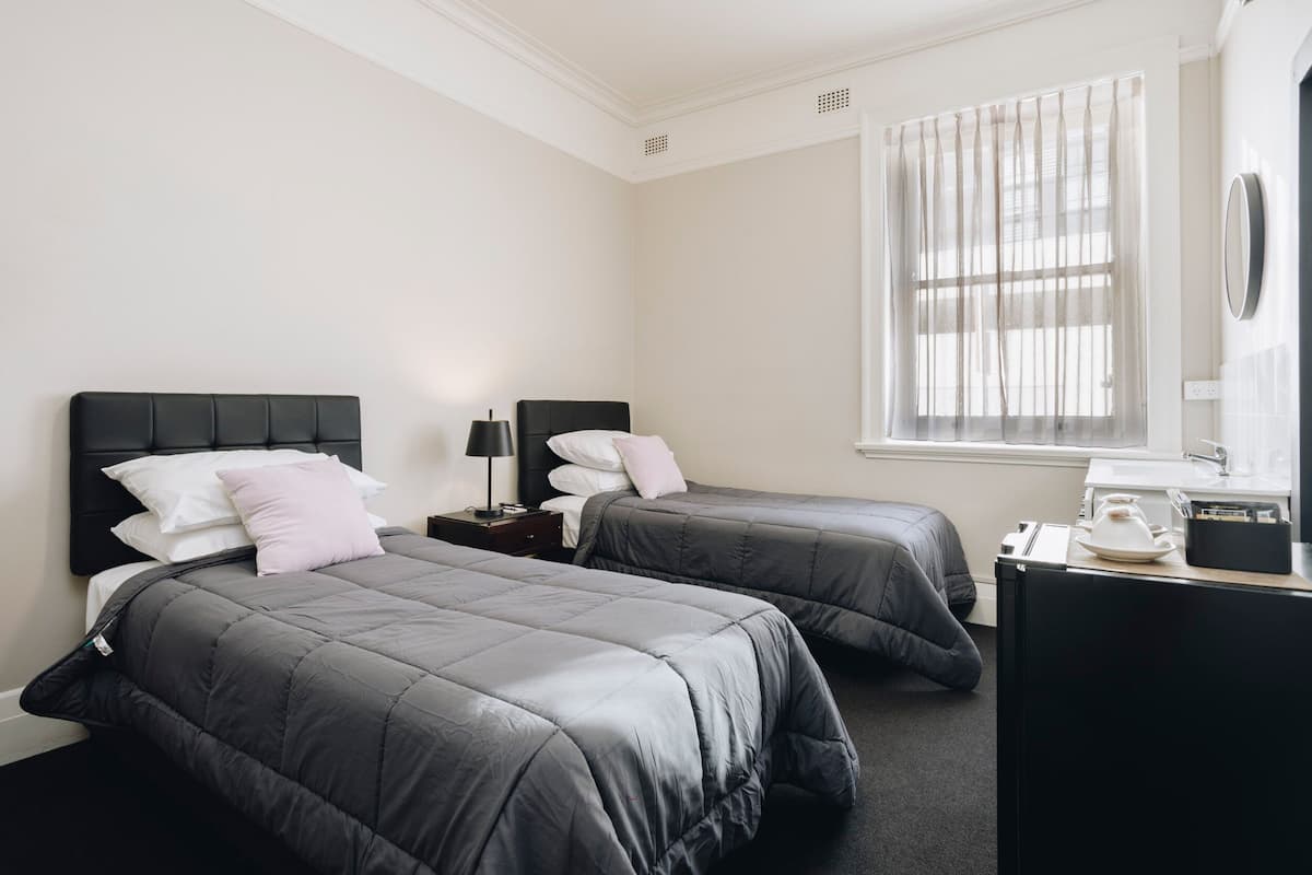 A hotel room with two beds, a desk, a window with sheer curtains, and a wall-mounted mirror.