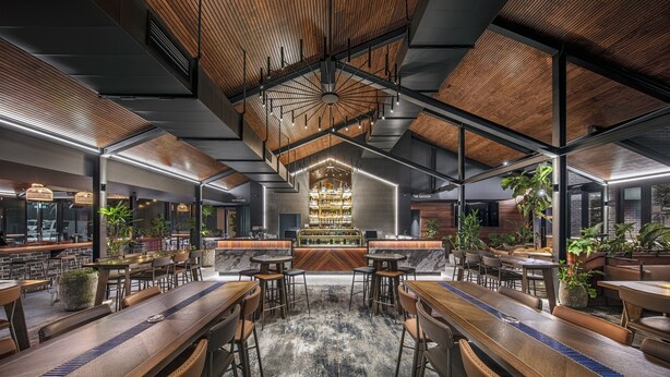 A modern bar area with a central bar counter, seating arrangements, and a ceiling design with exposed beams and lighting fixtures. - Hotel Gosford (Gosford)