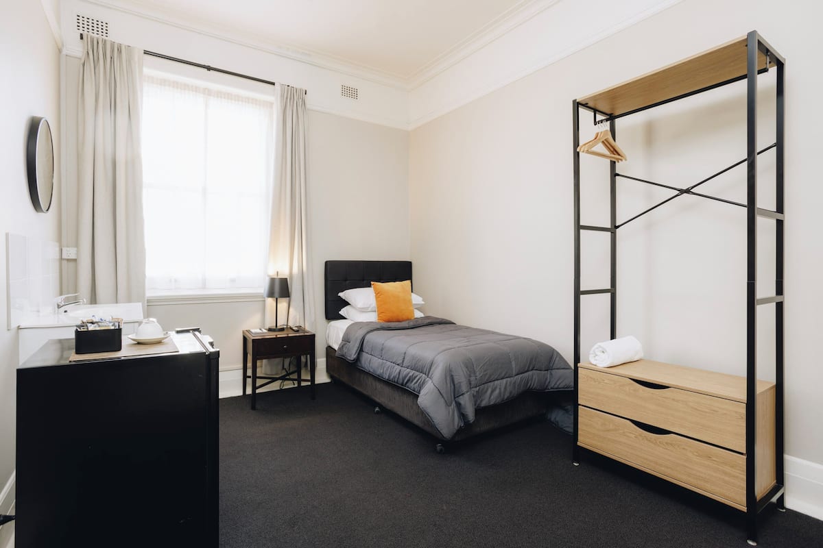 A compact hotel room with a bed, bedside table, dresser, and a built-in wardrobe with a mirror.