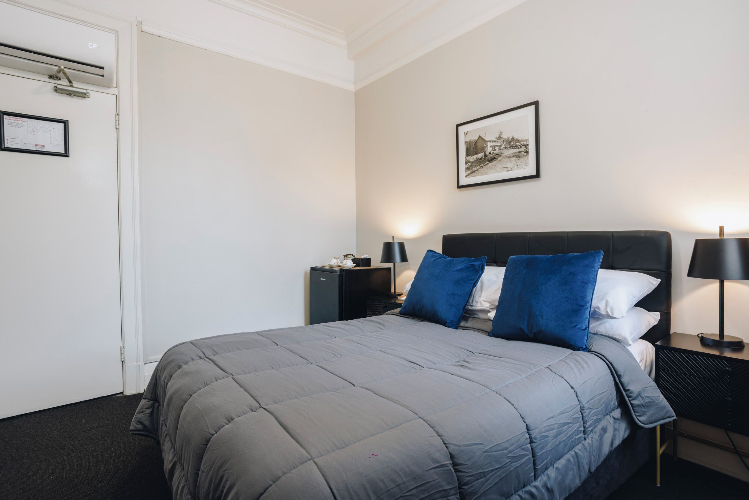 Un dormitorio ordenado con cama, mesitas de noche, una mesilla, un cuadro en la pared y una puerta con unidad de aire acondicionado.