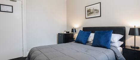 A neatly arranged bedroom with a bed, bedside tables, a nightstand, a framed picture on the wall, and a door with an air conditioning unit.