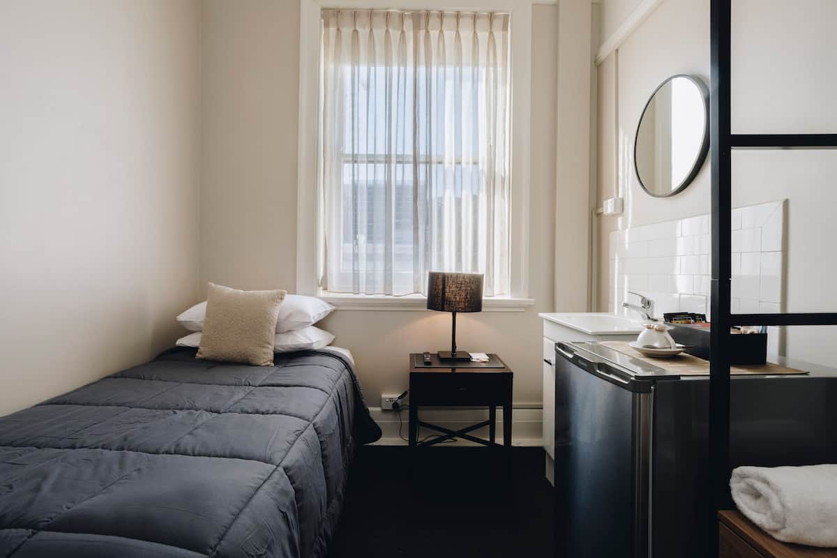 A small, neatly arranged bedroom with a bed, bedside lamp, and a window with sheer curtains.