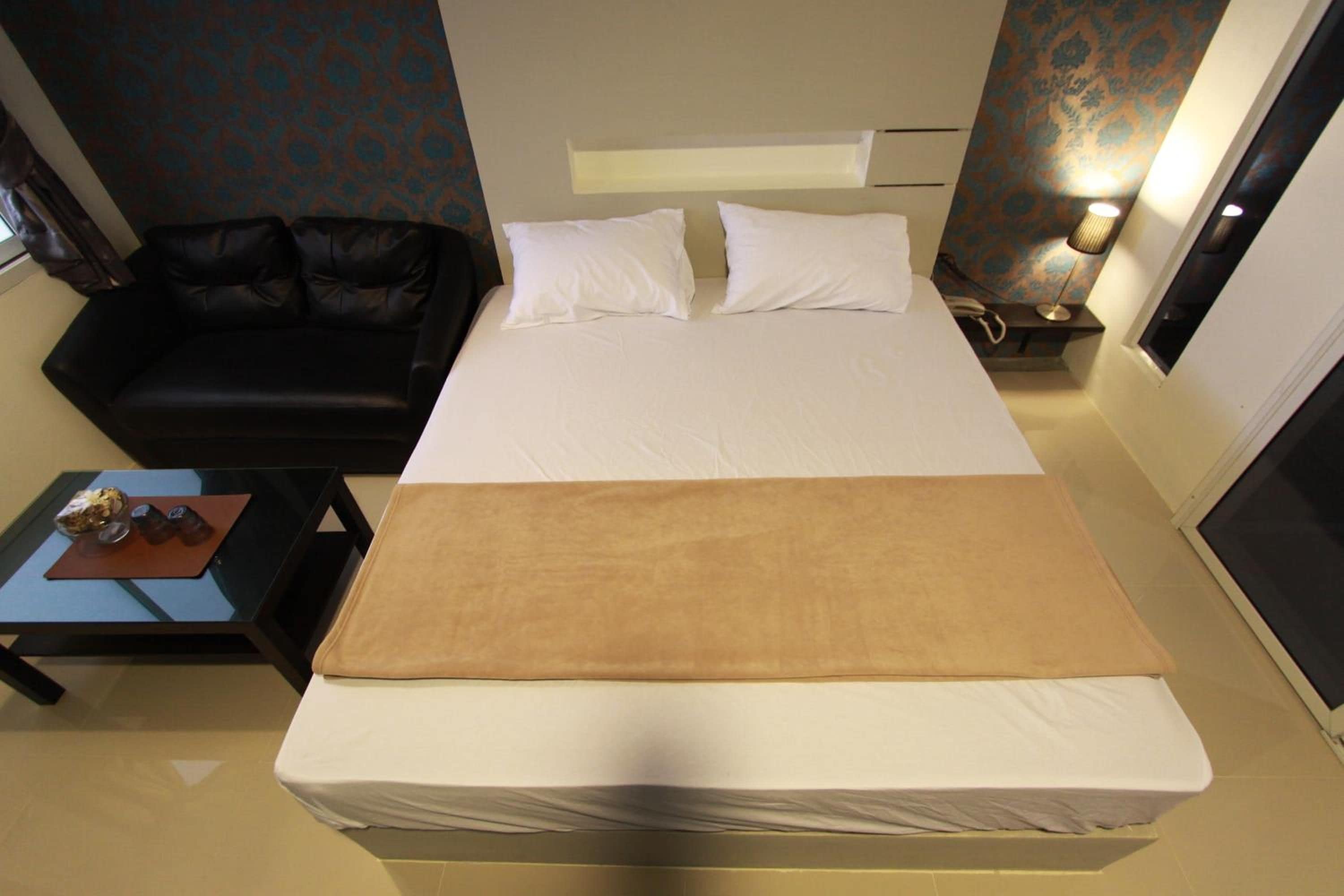 A hotel room with a bed, a black sofa, a small table with a bowl of cereal, and a lamp.