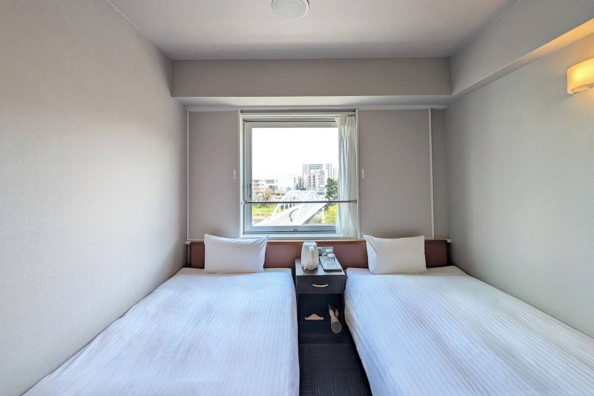 Een hotelkamer met twee bedden, een raam met uitzicht op gebouwen, een nachtkastje met een lamp, en een tafeltje met een telefoon en een glas.