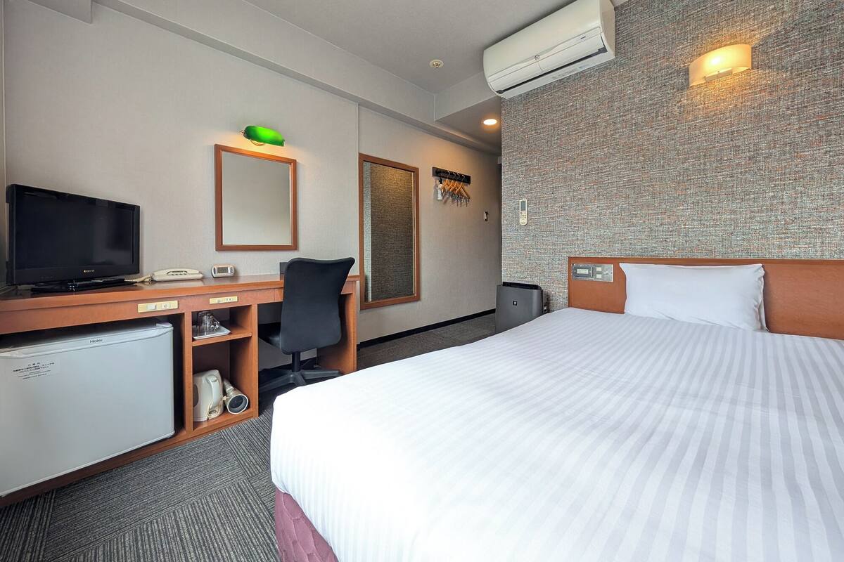 Een hotelkamer met een bed, een bureau met een computer, een televisie, een minibar en een wandairconditioner.