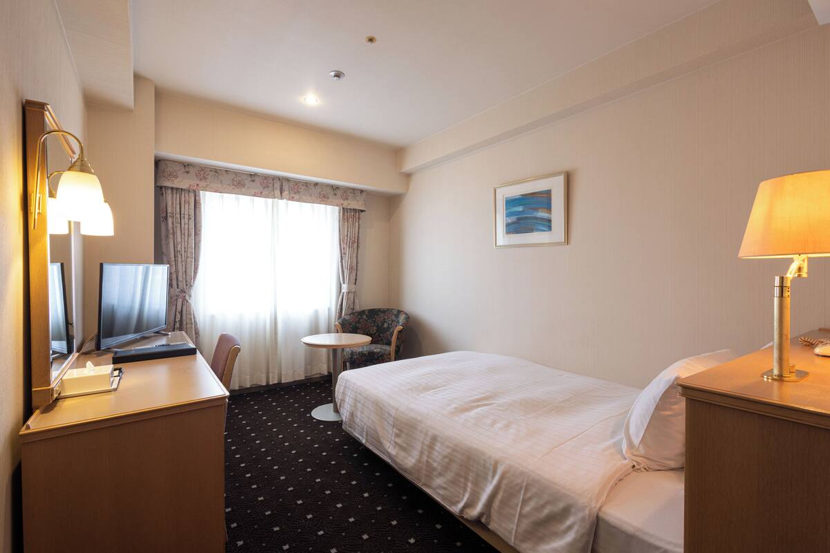 A hotel room with a bed, a desk with a computer, a chair, a small table, and a lamp.