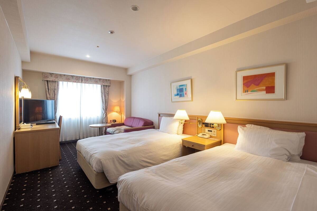 A hotel room with two beds, a desk with a television, a chair, a small table, and two lamps.