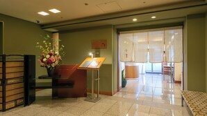 Un área de recepción con mostrador, un arreglo floral, un tablón de anuncios y una sala de espera con banca.