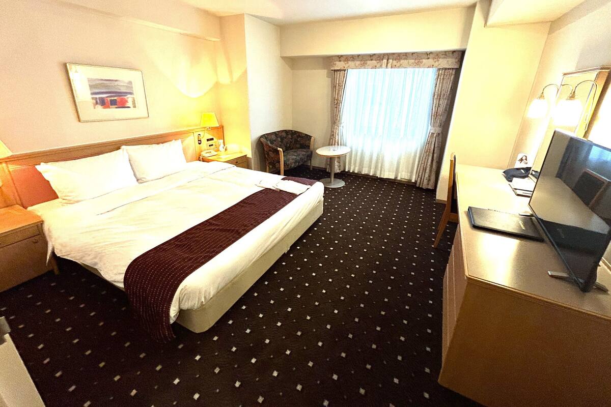 A hotel room with a bed, a desk with a laptop, a chair, a small table, and a window with curtains.