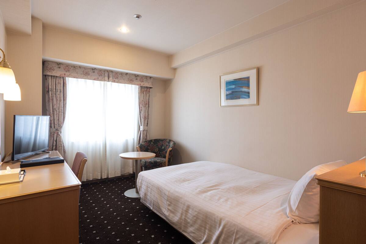 A hotel room with a bed, a desk with a computer, a chair, a small table, a lamp, a painting on the wall, and a window with curtains.