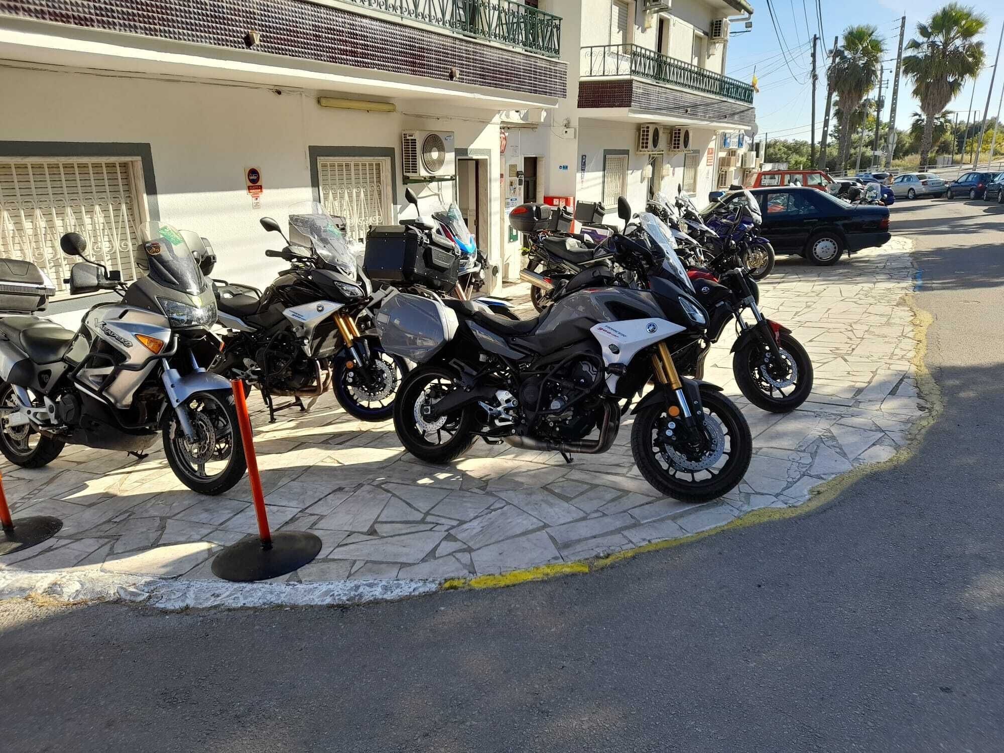 A row of parked motorcycles in front of a building with a tiled roof and balconies.