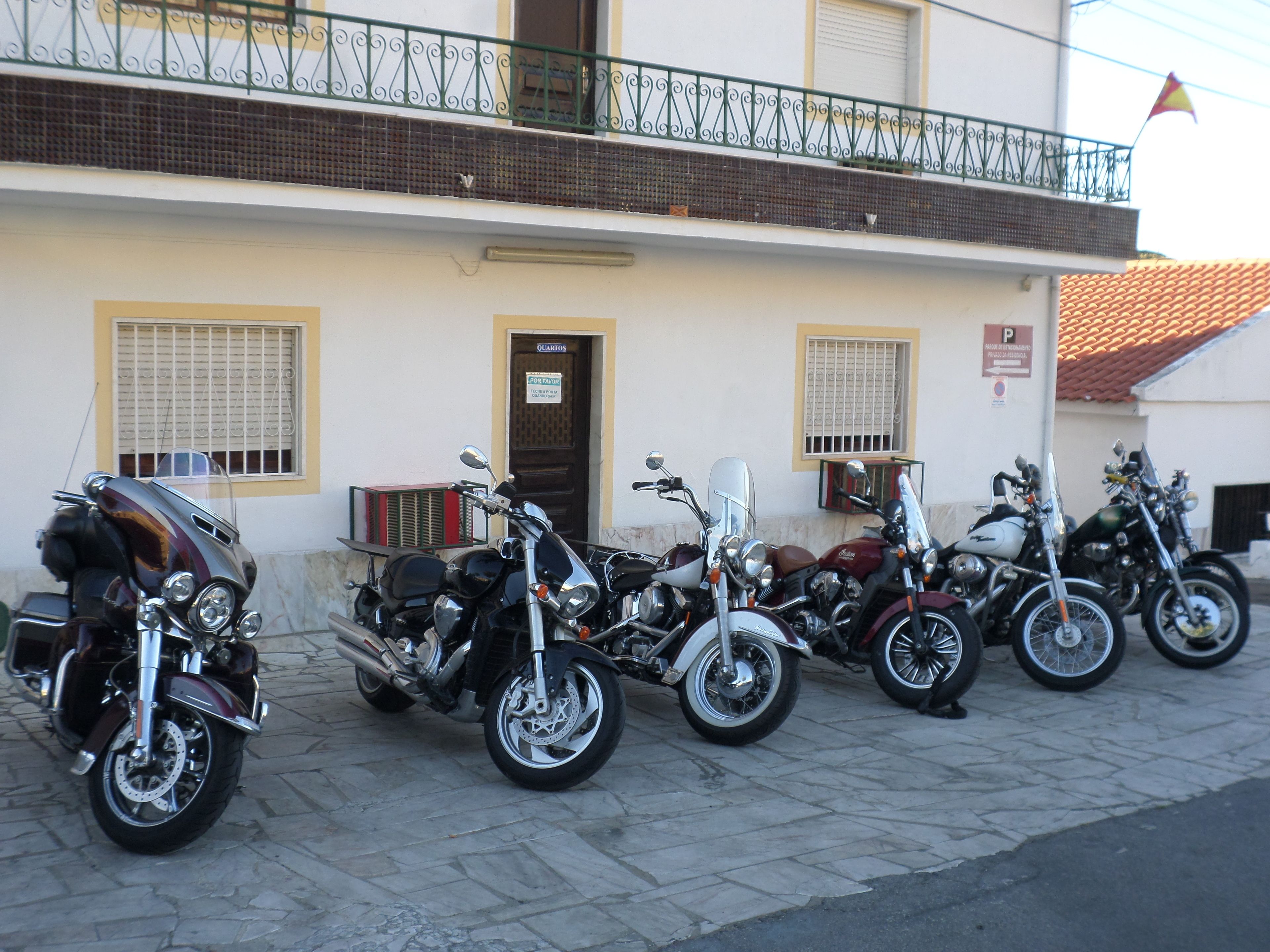 A row of motorcycles parked in front of a building with a tiled roof and a balcony with a metal railing.