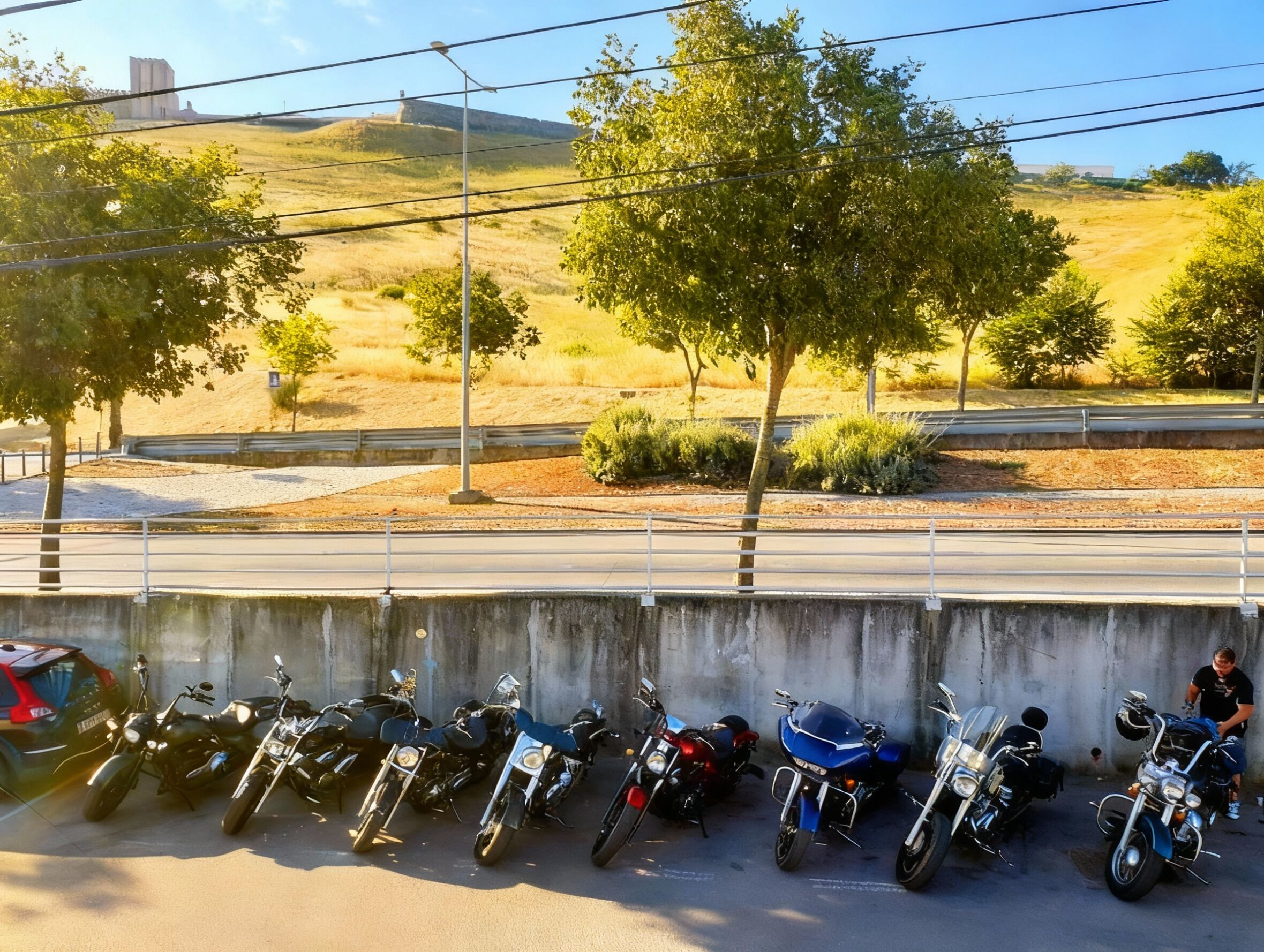 Una fila de motocicletas estacionadas al borde de una carretera, con una barrera de hormigón, árboles y un paisaje montañoso al fondo.