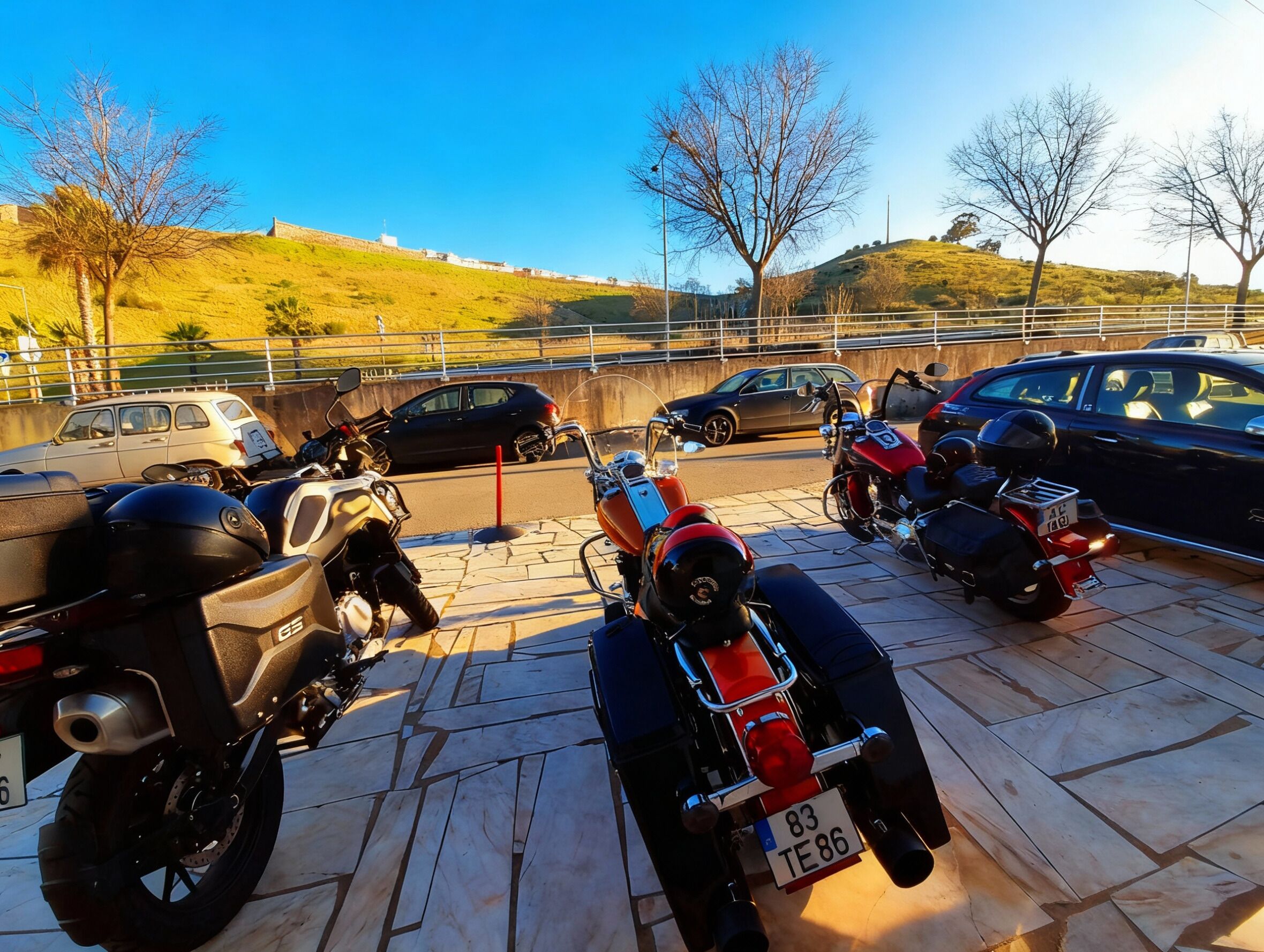 A parking area with motorcycles and cars, a clear sky, and a hilly landscape in the background.
