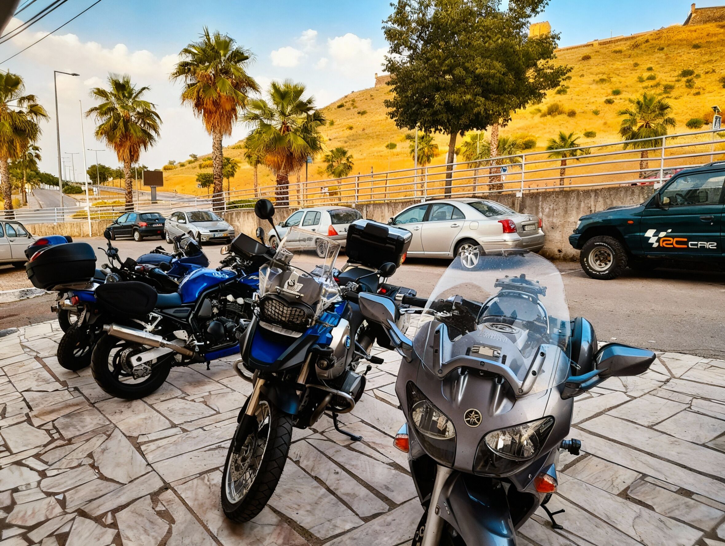 Un estacionamiento con varias motocicletas y automóviles, superficie empedrada de piedra y un fondo montañoso con vegetación escasa.
