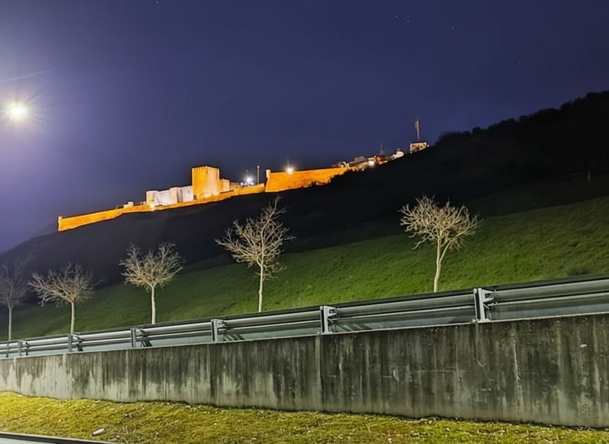 Ein Gebäude auf einem Hügel bei Nacht, beleuchtet, mit einer Betonbarriere im Vordergrund und vereinzelten Bäumen.