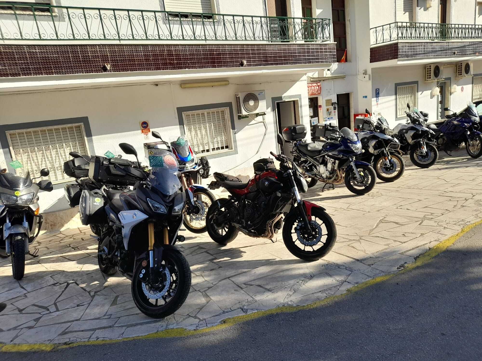 Fila di motociclette parcheggiate su un marciapiede asfaltato di fronte a un edificio con facciata piastrellata e un balcone con ringhiera in metallo.