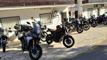 Fila di motociclette parcheggiate su un marciapiede asfaltato di fronte a un edificio con facciata piastrellata e un balcone con ringhiera in metallo.