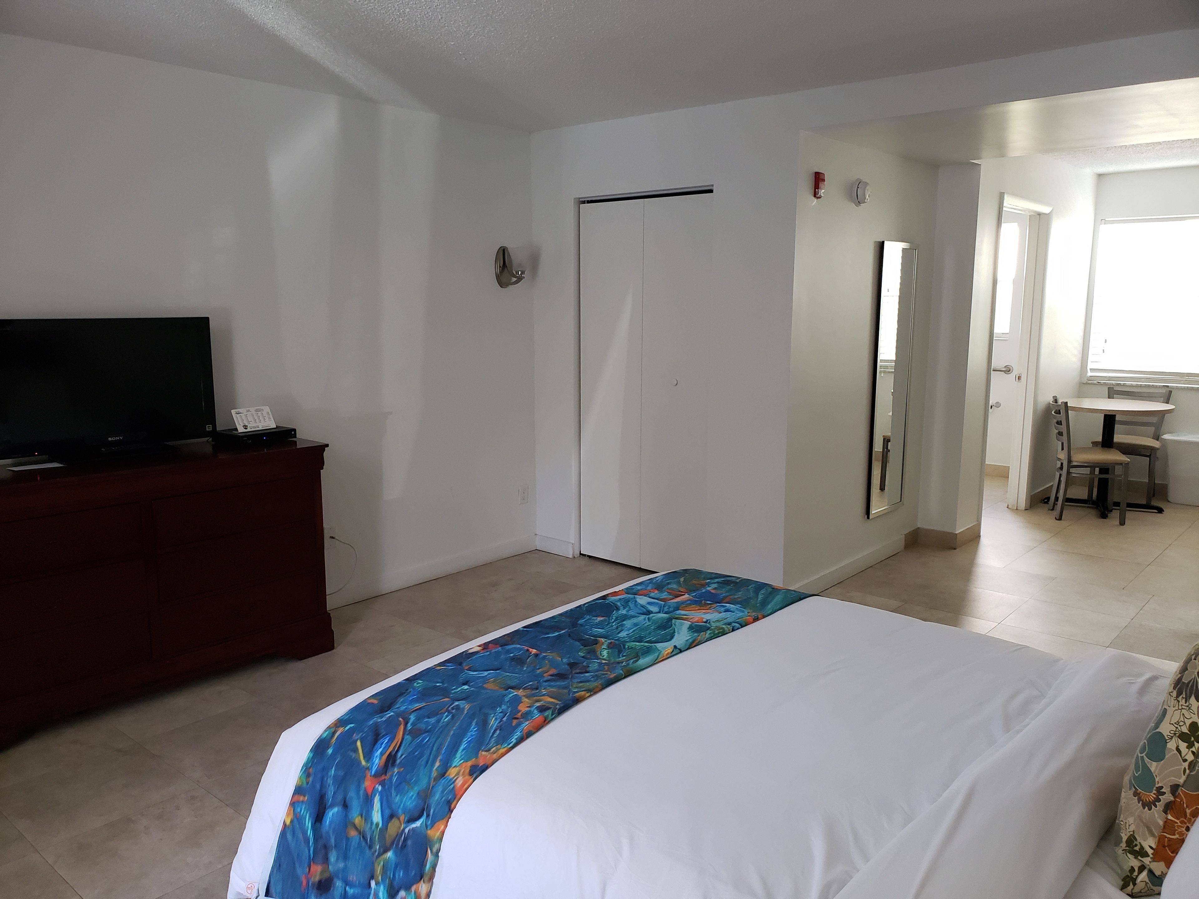 Habitación de hotel con una cama, un televisor sobre un aparador de madera, una puerta y una mesita con una silla.