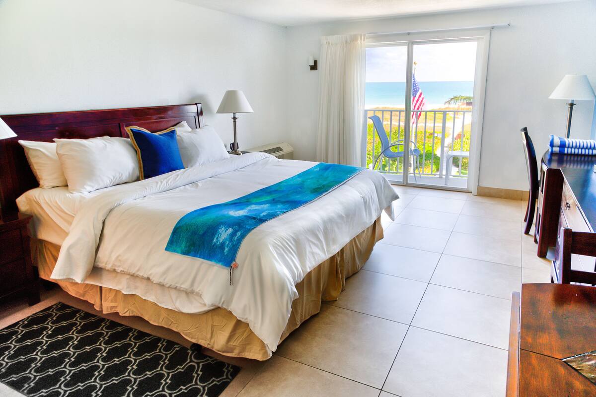 Ein Hotelzimmer mit einem großen Bett, einem Schreibtisch, einem Stuhl und einem Balkon mit Blick auf den Strand.