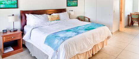 Habitación de hotel con una cama grande, una mesita de noche con lámpara, una silla y una puerta que da a un comedor.