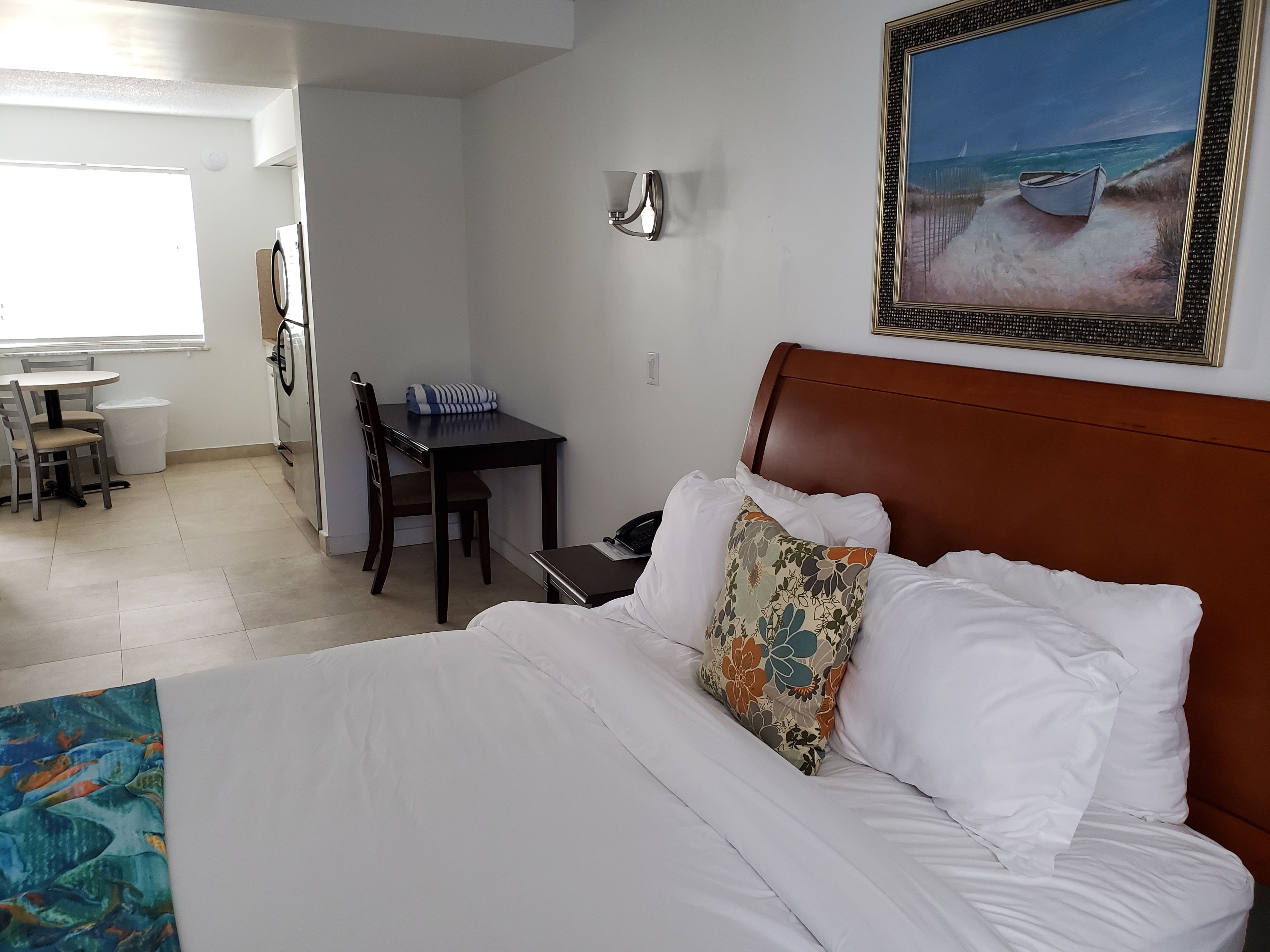 Una camera d'albergo con un letto, una scrivania, una sedia, una lampada e un quadro raffigurante una spiaggia.