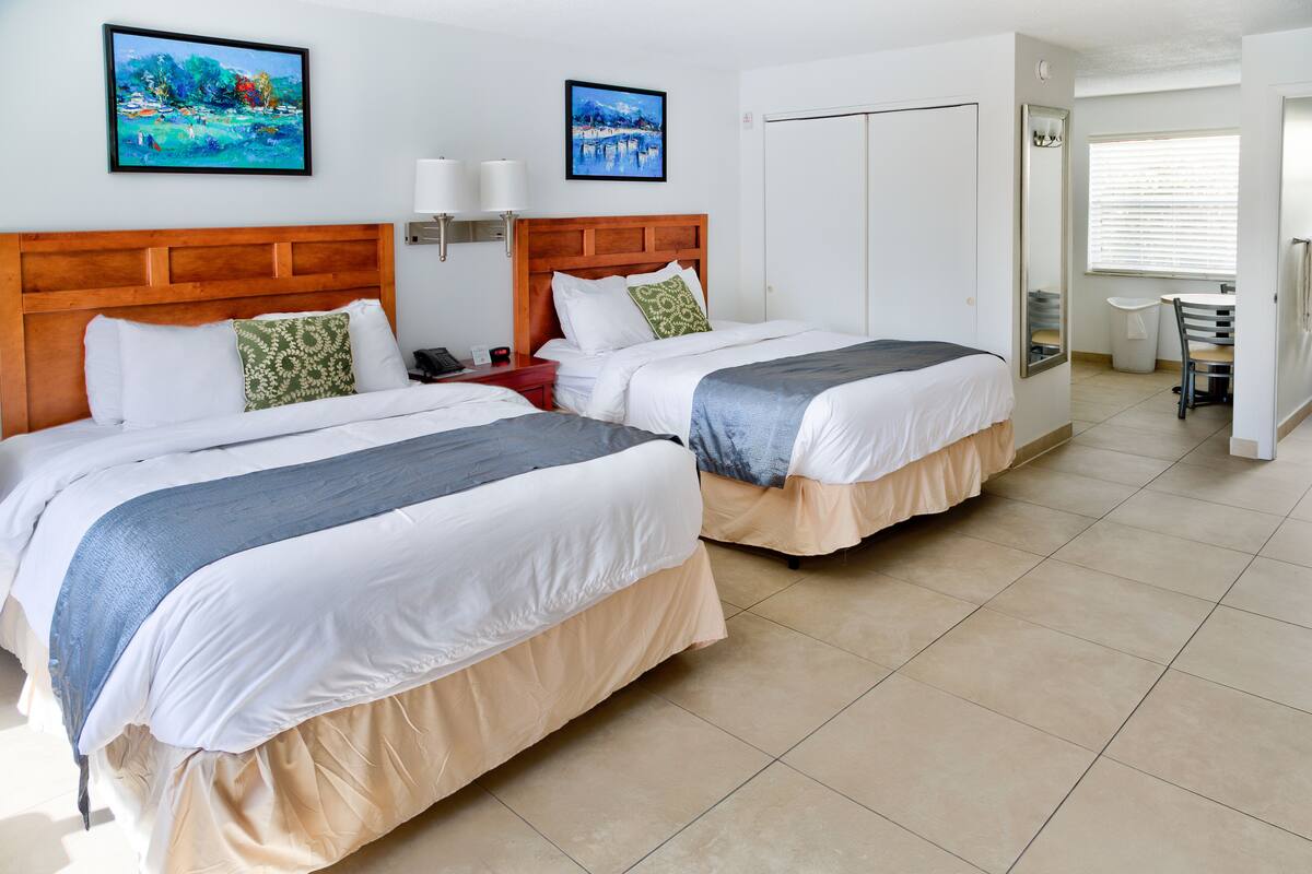 Ein Hotelzimmer mit zwei Betten, einem hölzernen Kopfteil, einem Nachttisch, einem Schreibtisch mit Stuhl, einem Fenster mit Jalousien und zwei Bildern an der Wand.