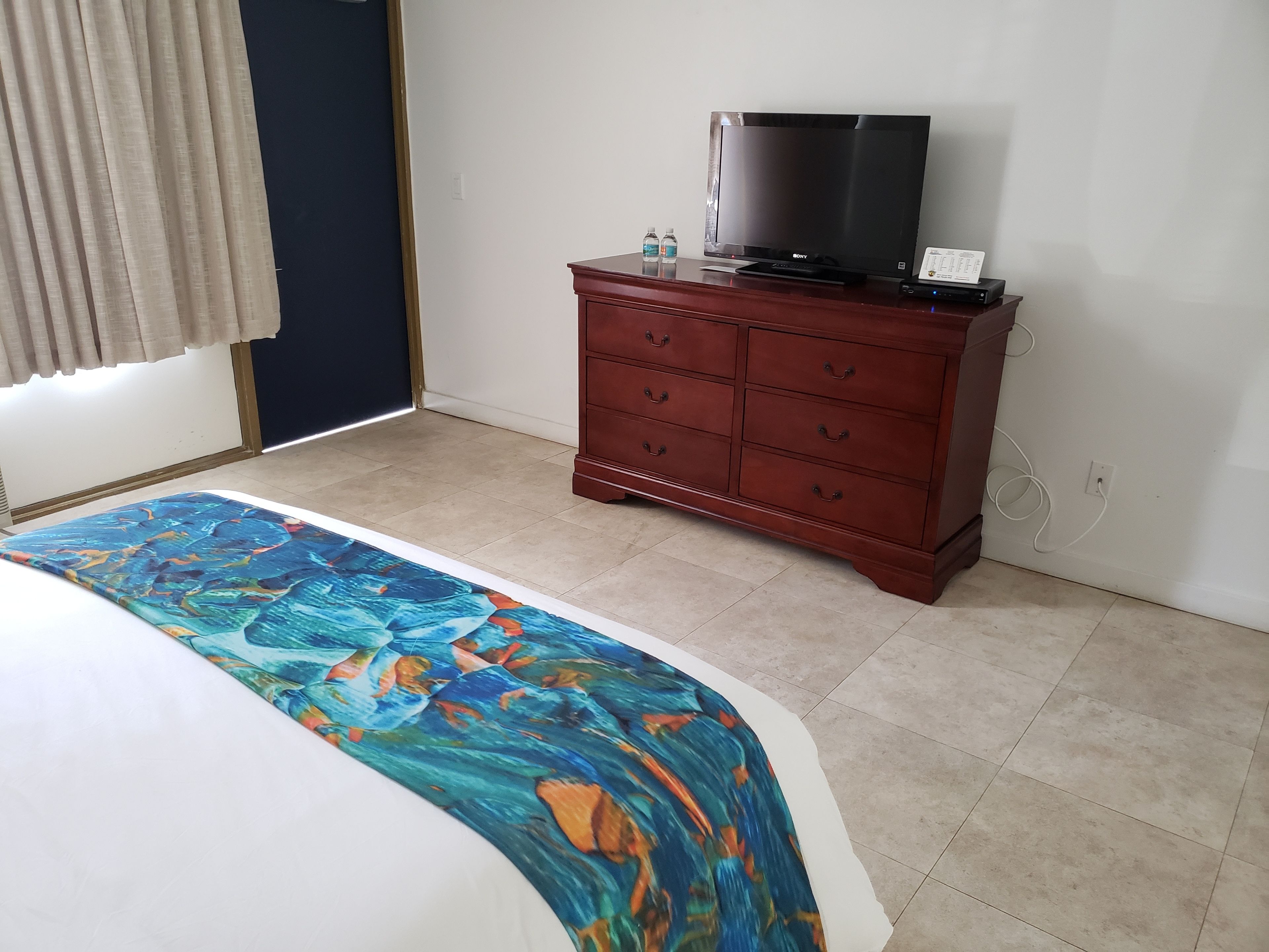 Une chambre d’hôtel avec un lit, une commode en bois, une télévision et une fenêtre avec des rideaux.