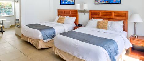 A hotel room with two beds, a desk, and a chair. There are two paintings on the wall, a window with blinds, and a lamp on the nightstand.