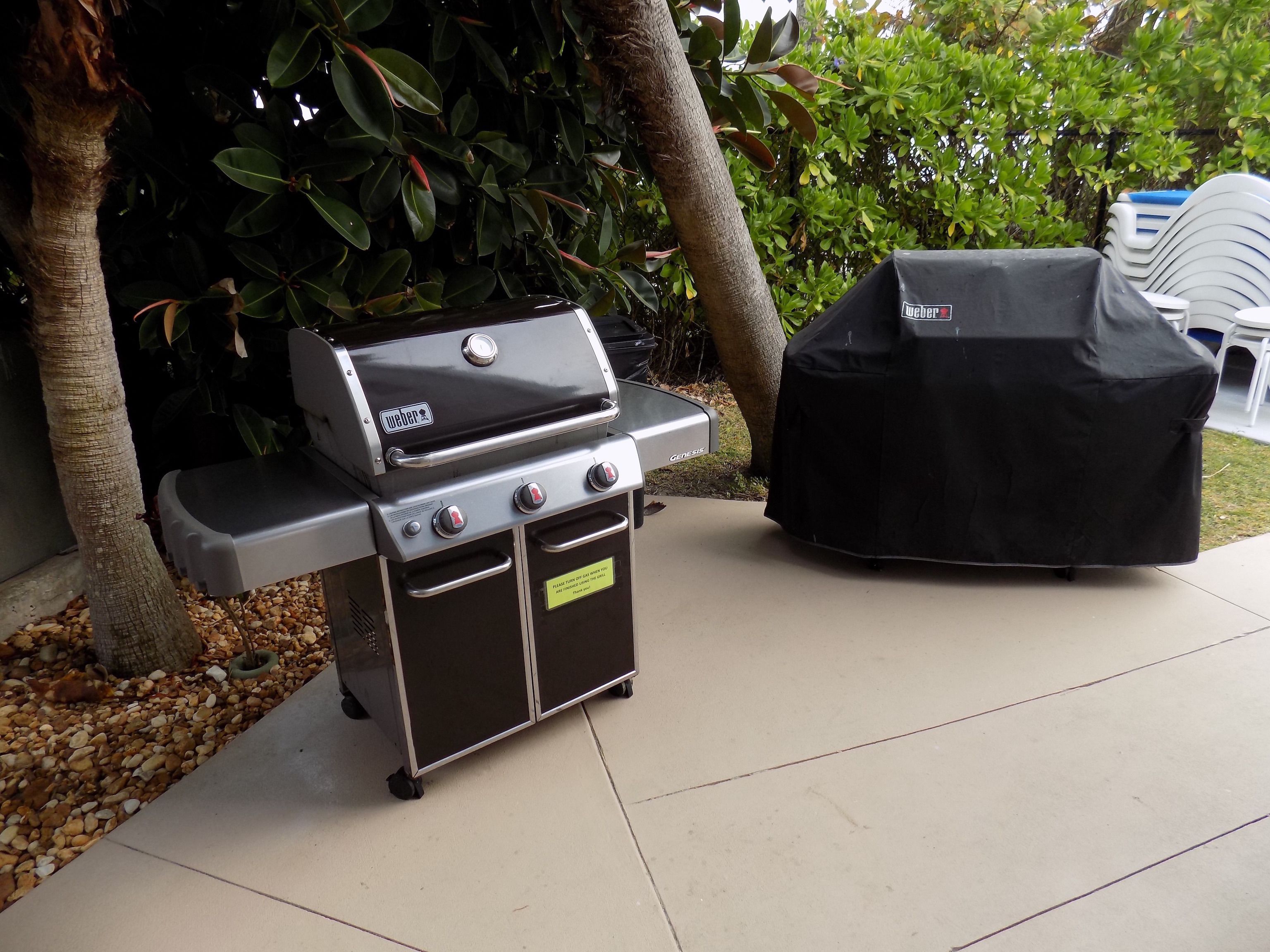 A portable Weber Genesis grill with a black cover and a side table, set on a patio with pebbles and greenery.