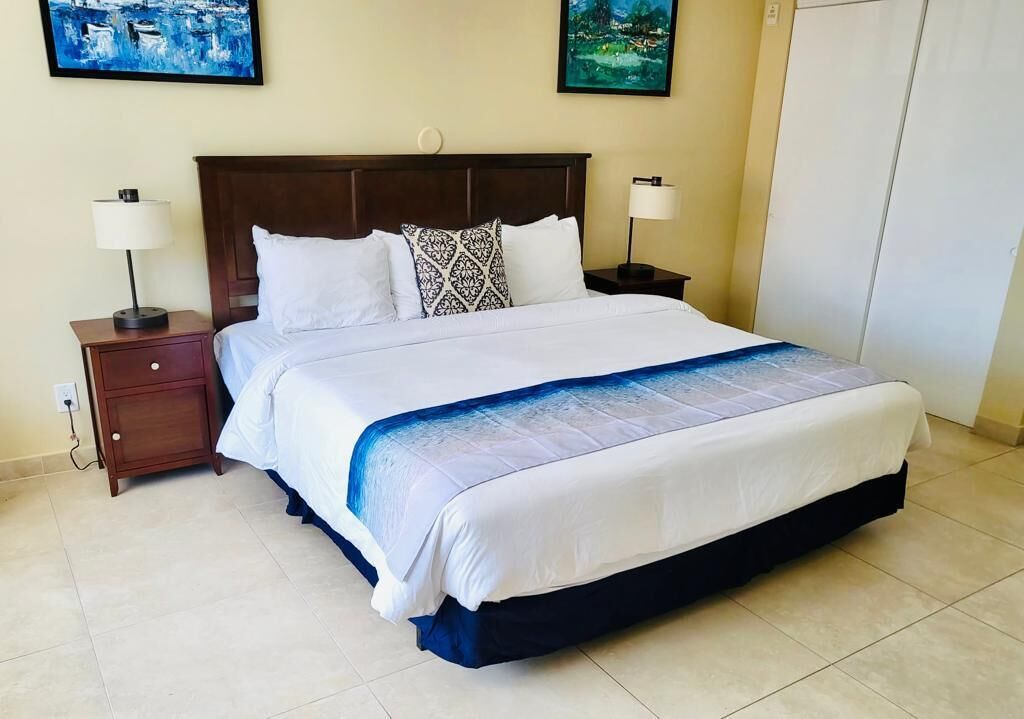 Una cama bien hecha con ropa de cama blanca y azul, dos mesitas de noche con lámparas y dos obras de arte enmarcadas en la pared.