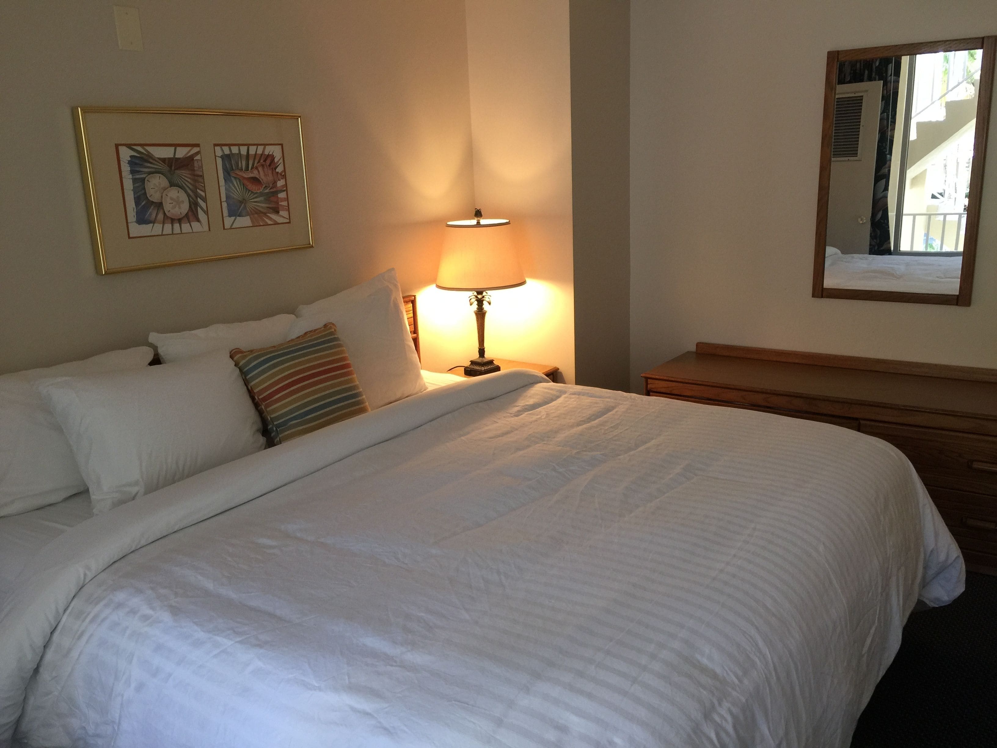 A neatly made bed with white linens and a striped pillow, a bedside lamp, and two framed artworks on the wall.