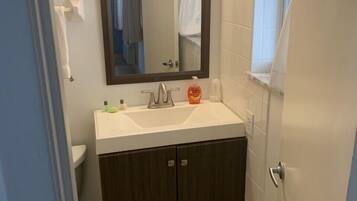 Un baño con lavamanos blanco, armarios de madera oscura, un espejo con luz superior y una toalla blanca colgada en un perchero.