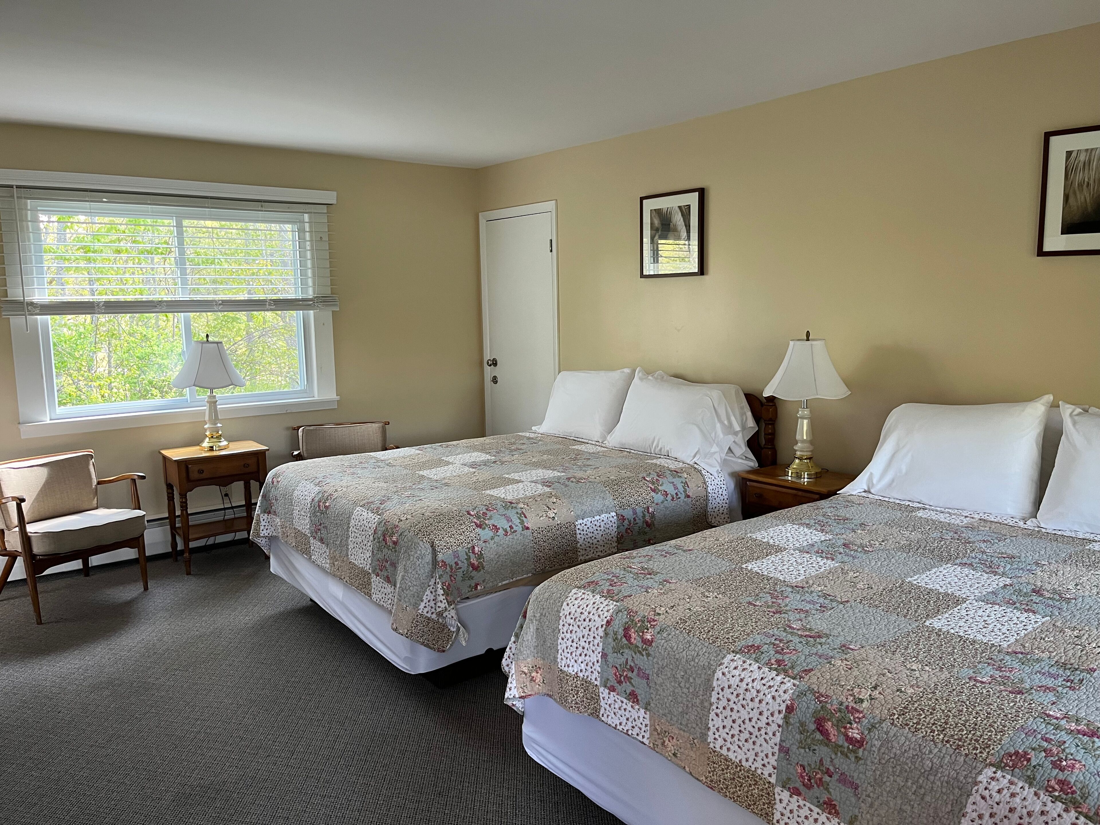 A hotel room with two beds, a desk, a chair, a window with blinds, and a door.