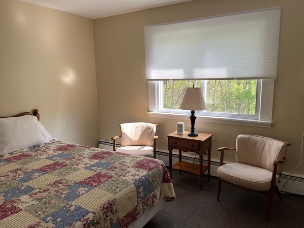 A bedroom with a bed, two chairs, a nightstand with a lamp, and a window with blinds.