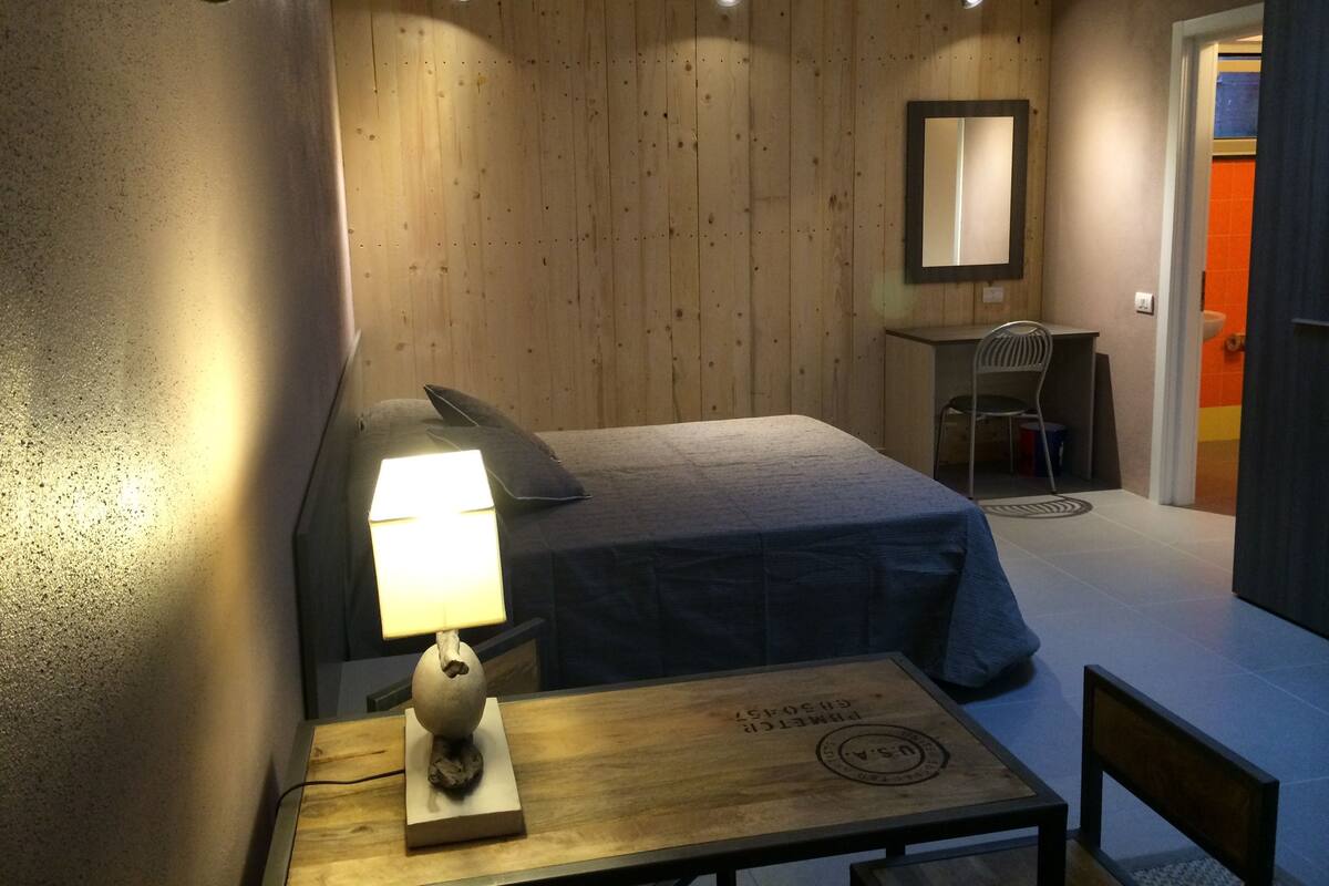 A room with a bed, a desk with a lamp, a chair, and a mirror on the wall.