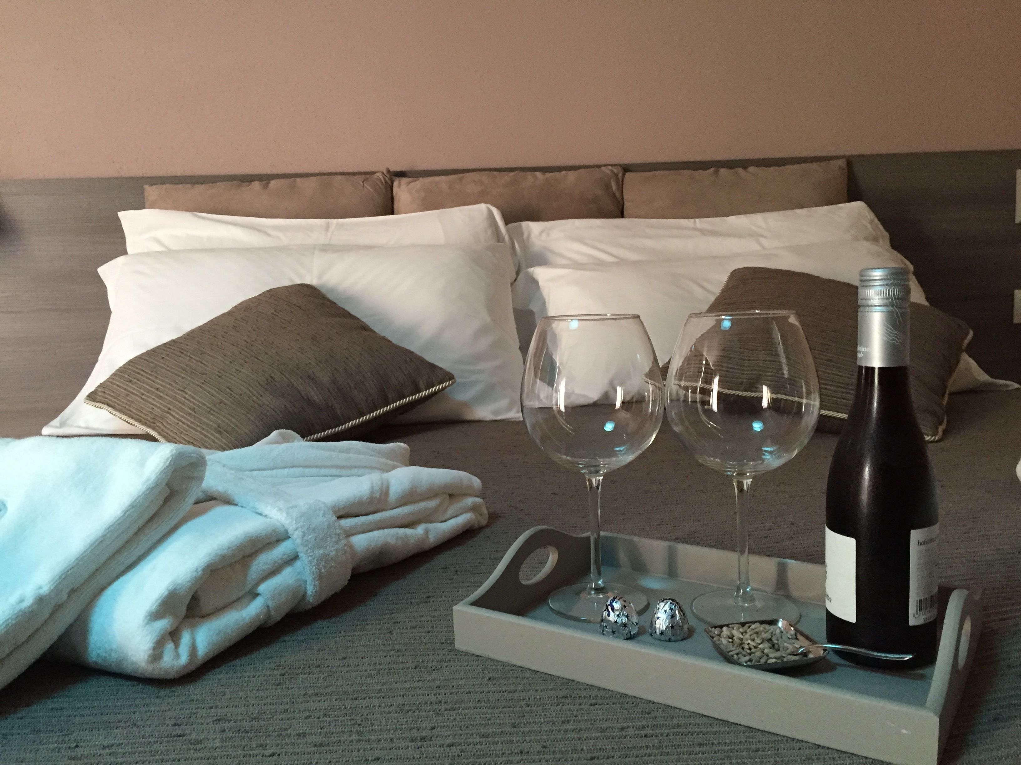 A neatly made bed with pillows, a folded towel, two wine glasses, a bottle of wine, and some chocolate on a tray.
