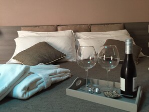 A neatly made bed with pillows, a folded towel, two wine glasses, a bottle of wine, and some chocolate on a tray. - Il Fortino Mini Suites (Ameglia)