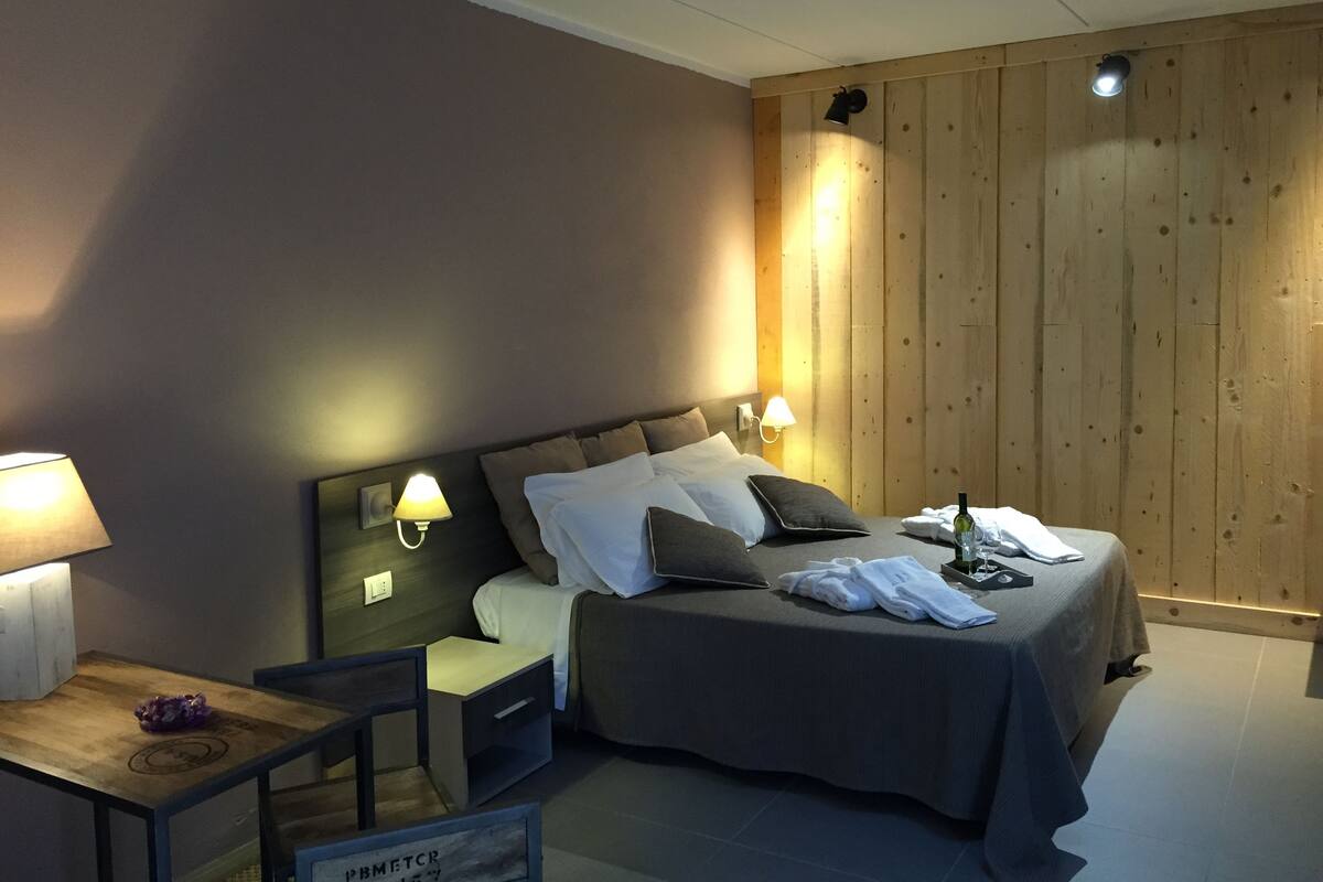 A hotel room with a bed, bedside lamps, a nightstand, a small table, and a wooden wall panel.