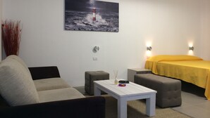 Una habitación con un sofá blanco, un reposapiés gris, una mesa blanca y una cama con cubrecolcha amarillo. En la pared, hay un cuadro de un faro.