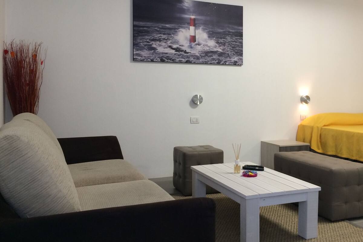 A room with a white sofa, a grey ottoman, a white table, and a bed with a yellow cover. There is a painting of a lighthouse on the wall.
