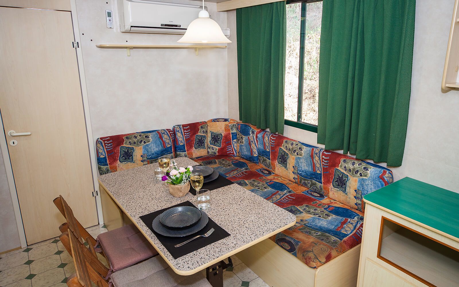 Un espacio habitable compacto con kitchenette, un área de comedor con mesa y banco, y una ventana con cortinas verdes.