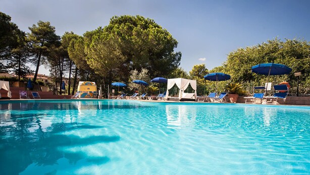 Una piscina azul cristalina rodeada de sillas de descanso y sombrillas, con árboles y un edificio al fondo.