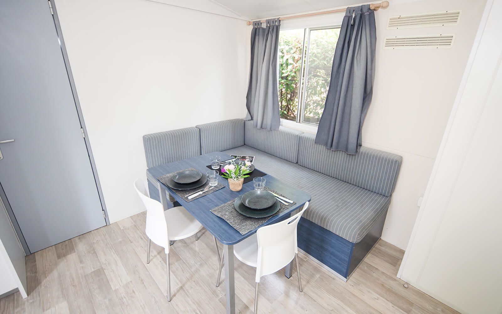 A compact room with a dining table set for two, a grey-striped sofa, and a window with curtains.